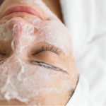 Rosto de pessoa com olhos fechados coberto por espuma branca de limpeza facial, sugerindo um tratamento de pele relaxante.