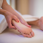 Pés apoiados sobre uma toalha branca enquanto mãos aplicam creme e massageiam suavemente, em um ambiente de spa para cuidados com os pés.